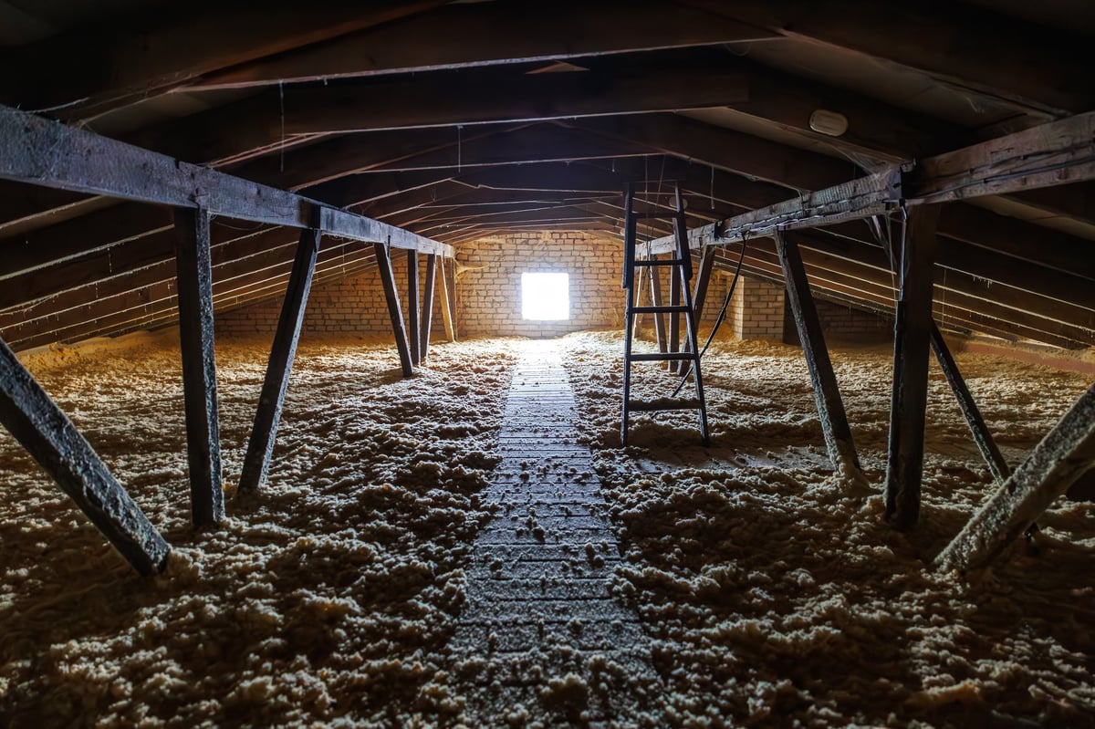 A long attic shows loose blown in insulation, a plank walkway to a bright square window, a metal step ladder, side wires, and rough beams in warm late day light.